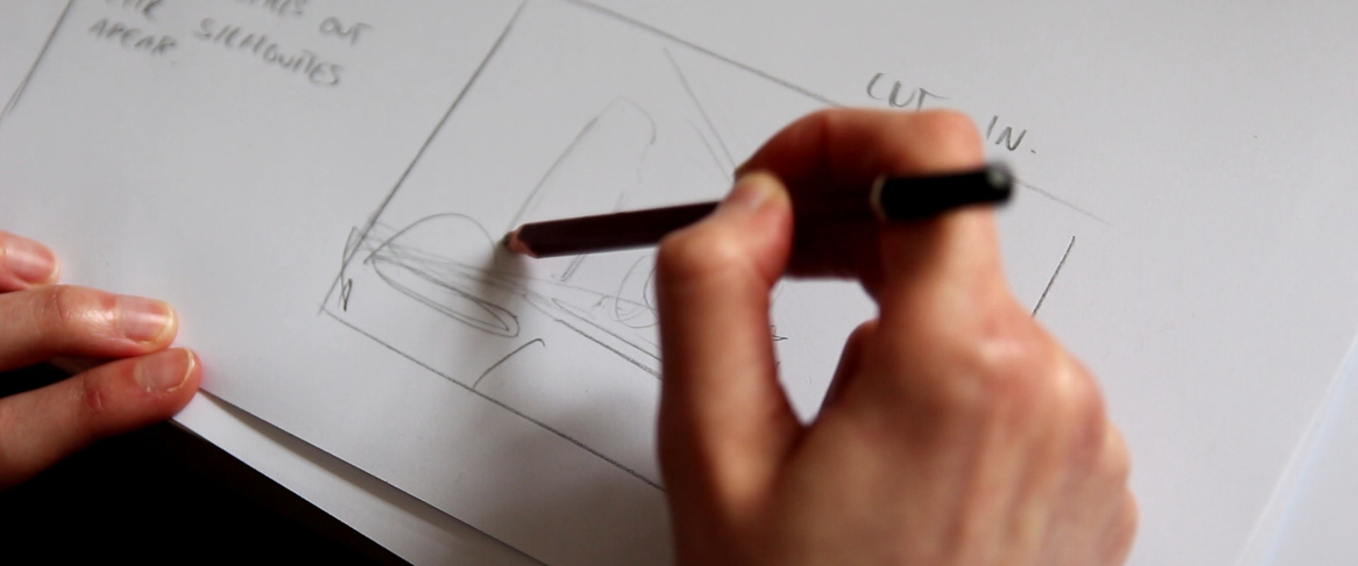 A hand drawing a storyboard on white paper with a pencil